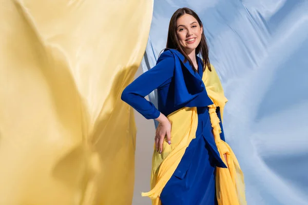 happy ukrainian woman in color block clothing posing with hand on hip near blue and yellow fabric