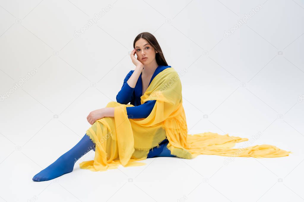 dreamy ukrainian young woman in blue and yellow outfit with tights sitting while posing on grey