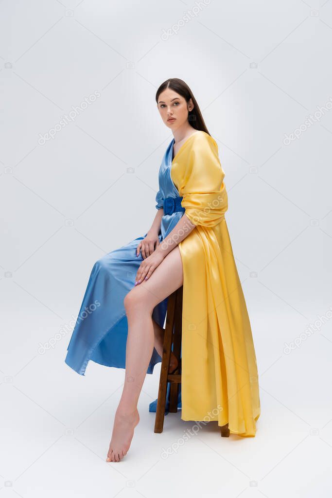 full length of sensual young ukrainian woman in blue and yellow dress posing while sitting on grey