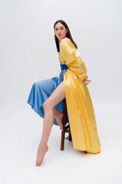 barefoot young ukrainian woman in blue and yellow dress posing while sitting on wooden chair on grey