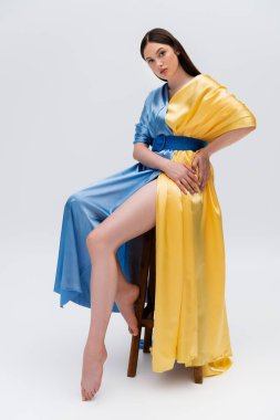 barefoot ukrainian woman in blue and yellow dress posing while sitting on wooden chair on grey