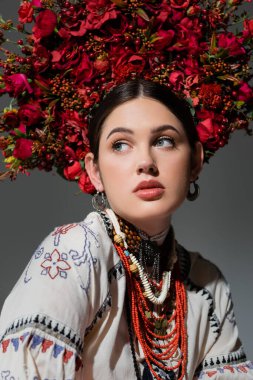 portrait of ukrainian woman in traditional clothing and floral red wreath looking away isolated on grey