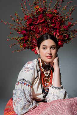 portrait of young ukrainian woman in traditional costume and floral red wreath isolated on grey