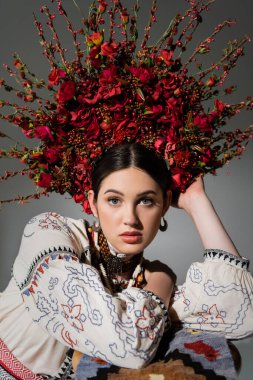 portrait of ukrainian woman in traditional clothes and floral wreath with red berries looking at camera isolated on grey clipart