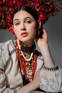 portrait of pretty ukrainian woman in traditional clothes adjusting floral red wreath isolated on grey