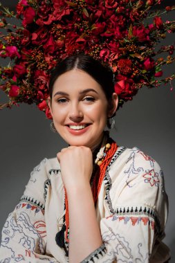 portrait of smiling ukrainian woman in traditional clothes and red wreath with flowers and berries isolated on grey
