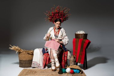 full length of ukrainian woman in traditional clothes and red floral wreath holding knitting needles on dark grey