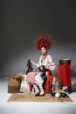 full length of young ukrainian woman in traditional clothes and floral wreath with berries holding knitted scarf on dark grey