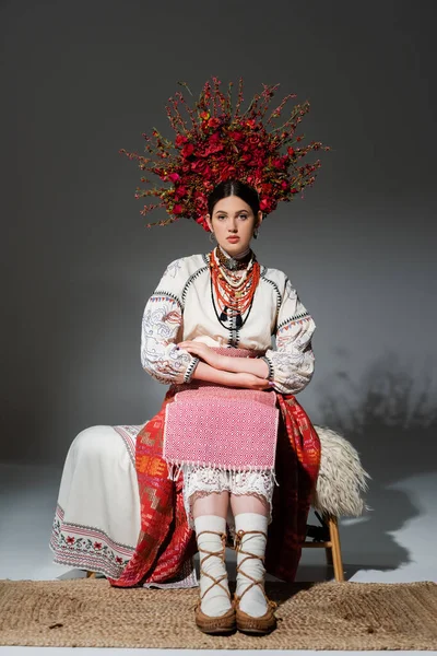 full length of young ukrainian woman in traditional clothing and floral red wreath sitting on bench on grey