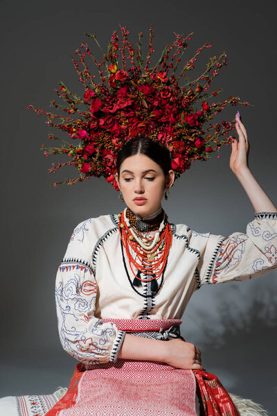 portrait of young ukrainian woman in traditional clothing and floral red wreath on grey