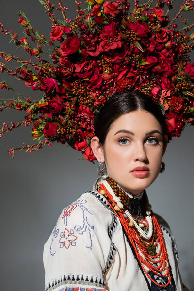 portrait of pretty ukrainian woman in traditional clothing and floral red wreath isolated on grey