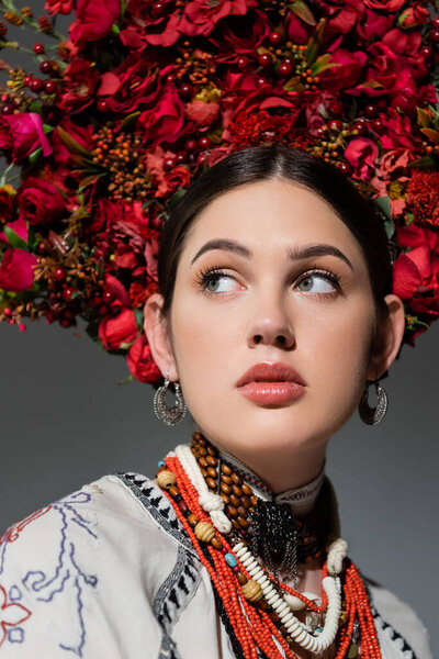 portrait of pretty ukrainian woman in traditional clothing and floral red wreath looking away isolated on grey