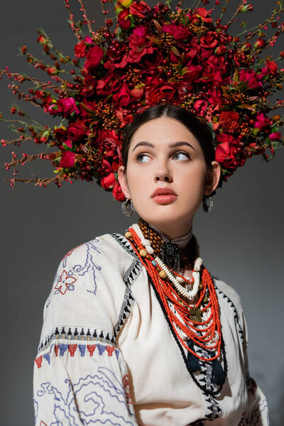 portrait of brunette ukrainian woman in traditional clothing and floral red wreath looking away isolated on grey