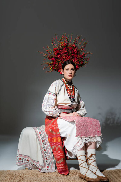 full length of pretty ukrainian woman in traditional clothing and red wreath with flowers and berries on grey