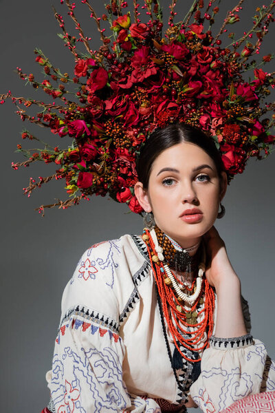 portrait of pretty ukrainian woman in traditional clothing and floral red wreath with berries isolated on grey