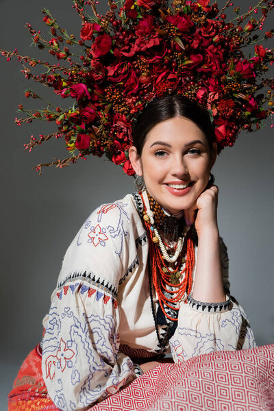 portrait of happy ukrainian woman in traditional clothing and floral red wreath isolated on grey