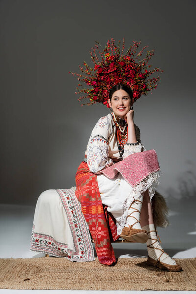 full length of happy ukrainian woman in traditional clothes and red wreath with flowers and berries on grey