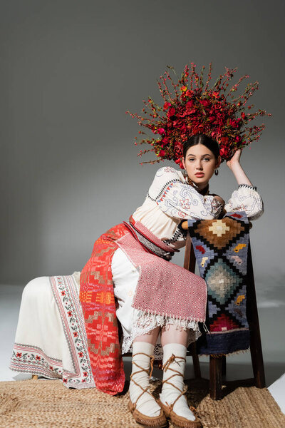 full length of pretty ukrainian woman in traditional dress and red wreath with flowers and berries on grey