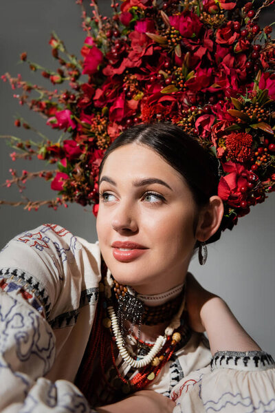 portrait of smiling ukrainian woman in traditional clothes and floral wreath with red berries isolated on grey