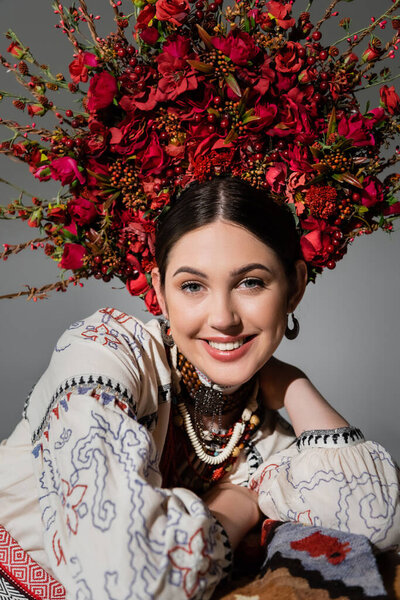 portrait of joyful ukrainian woman in traditional clothes and floral red wreath isolated on grey