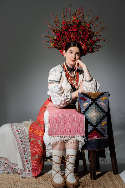 thoughtful ukrainian woman in traditional clothes and red wreath with flowers and berries on grey