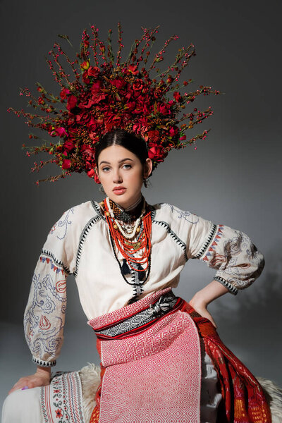 pretty ukrainian woman in traditional clothes and red wreath with flowers posing with hand on hip on grey