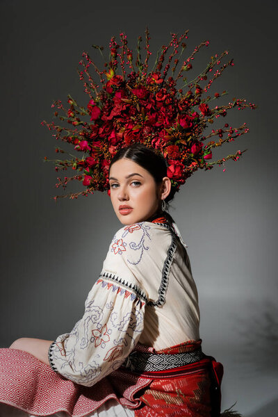 portrait of pretty ukrainian woman in traditional costume and floral red wreath on grey
