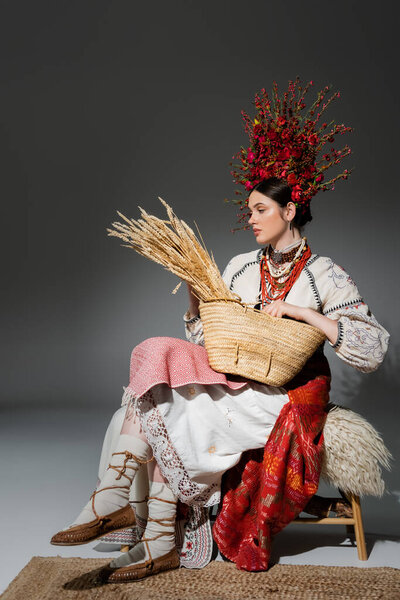 full length of pretty ukrainian woman in traditional clothes and red wreath with flowers holding bag with wheat on dark grey