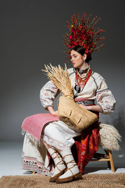 full length of young ukrainian woman in red wreath with flowers holding bag with wheat on dark grey