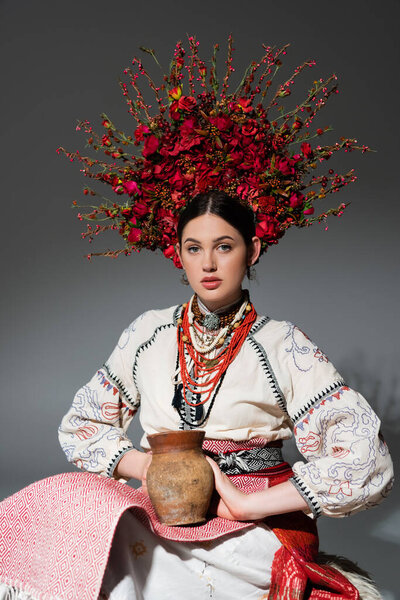 young ukrainian woman in traditional clothes and red wreath with flowers holding clay pot on grey