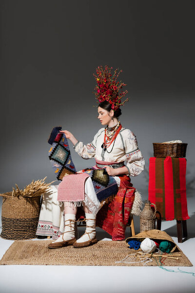 full length of young ukrainian woman in traditional clothes and red floral wreath holding knitted scarf on dark grey