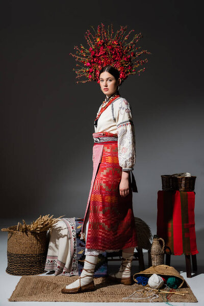 full length of ukrainian woman in traditional red floral wreath standing near yarn balls and wheat on dark grey