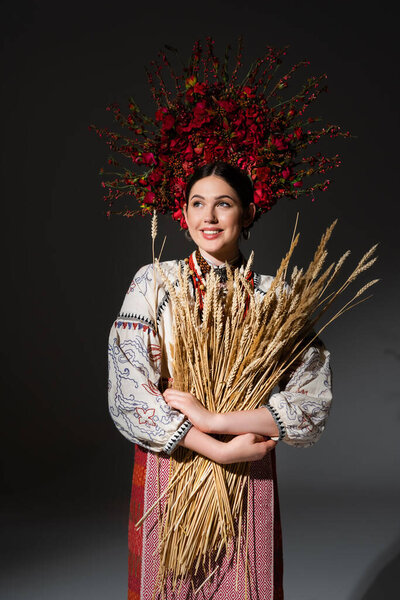 happy and young ukrainan woman in floral wreath with red berries holding wheat spikelets on black