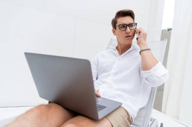 Gözlüklü bir adam dizüstü bilgisayarla güvertede oturuyor ve cep telefonuyla konuşuyor. 