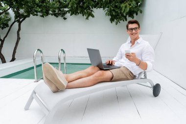 full length of joyful man with gadgets looking away while sitting in deck chair near pool
