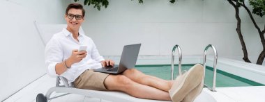 full length of cheerful man with mobile phone and laptop sitting in deck chair near pool, banner