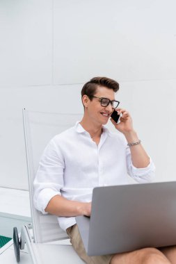 Gözlüklü mutlu adam dizüstü bilgisayarla güvertede oturuyor ve akıllı telefondan konuşuyor.