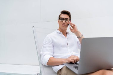 Gözlüklü serbest çalışan güverte koltuğunda oturuyor ve cep telefonuyla dizüstü bilgisayarın yanında konuşuyor.