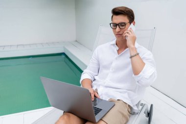 man in eyeglasses sitting in deck chair with laptop and talking on smartphone near pool