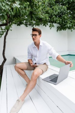 full length of man with laptop and mobile phone sitting near pool under green branches