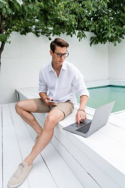 freelancer in eyeglasses sitting at poolside with mobile phone and using laptop