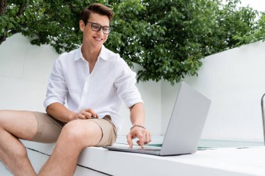 happy man in summer clothes sitting at poolside and looking at laptop