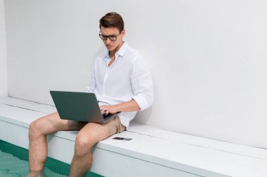 man in eyeglasses tying on laptop while sitting at poolside near mobile phone