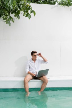 man in white shirt sitting near pool with laptop and looking away