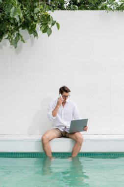 man sitting with feet in pool and talking on mobile phone near laptop