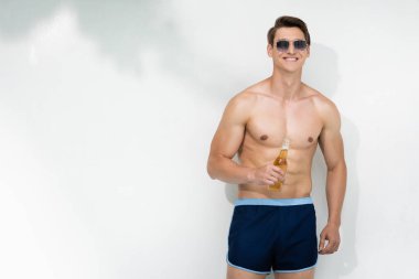 Güneş gözlüğü ve mayoyla beyaz duvarın yanında dikilen mutlu adam elinde bira şişesi tutuyor.