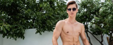 Açık havada yeşil dalların yanında güneş gözlüklü üstsüz sporcu.