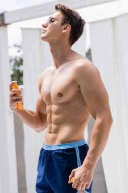 shirtless athletic man in sunglasses applying sunscreen on body