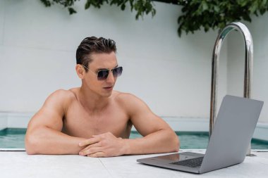 shirtless man in sunglasses looking at laptop near pool