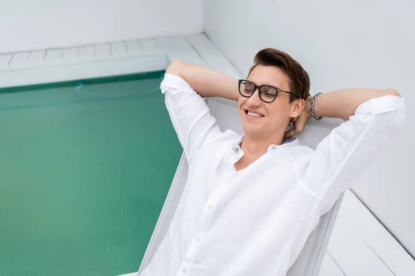 happy man in eyeglasses and silver bracelet resting in deck chair with hands behind head